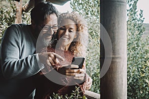 Happy couple using mobile phone having fun together outside on the terrace at home. Man and woman smile and use cellular to video