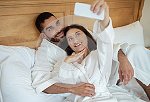 Happy Couple Using Smartphone Making Selfie Posing Lying In Bedroom