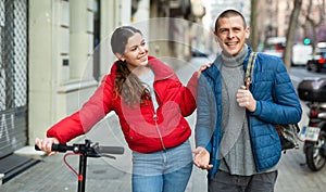 Couple are walking along the spring street. Girl with electric scooter