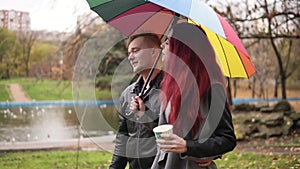 Happy couple walking together in autumn park holding a colorful umbrella. Attractive man in leather jacket is embracing