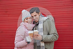 Happy couple in warm clothes with drinks near red wall outdoors. Christmas season