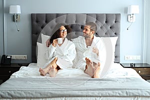 Happy couple in white bathrobe with cup of coffee on a bed in a hotel.