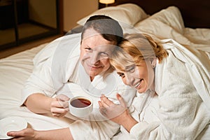 Happy couple in white bathrobes relaxing in bed