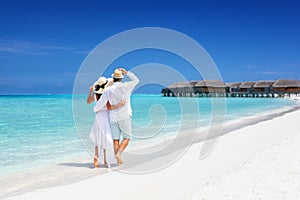 Happy couple in white clothing and with hats walks down a tropical beach