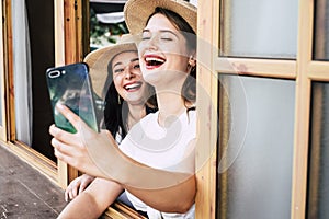 Happy couple of young woman caucasian friends have fun together at the window enjoying the phone internet connection - cheerful