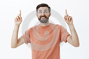 Happy curious attractive european man with dark beard in casual t-shirt raising index fingers looking and pointing up