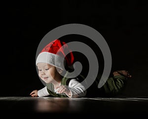 Happy curious infant boy toddler in red Santa hat and white - green clothes is lying on floor looking at copy space