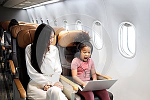 Happy curly hair African girl child passenger with headphones using laptop computer, sitting in seat inside airplane, cheerful kid