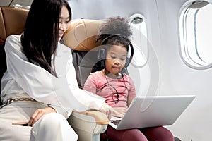 Happy curly hair African girl child passenger with headphones using laptop computer, sitting in seat inside airplane, cheerful kid