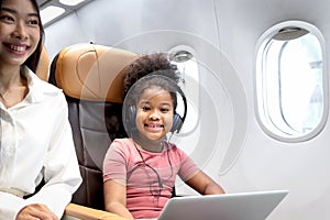 Happy curly hair African girl child passenger with headphones using laptop computer, sitting in seat inside airplane, cheerful kid