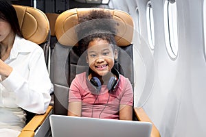 Happy curly hair African girl child passenger with headphones using laptop computer, sitting in seat inside airplane, cheerful kid