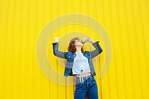 Happy curly haired woman in denim clothes drinking coffee on yellow background.