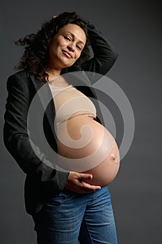 Happy curly multi-ethnic pregnant woman gently holding her naked belly, smiling looking at camera, isolated on gray