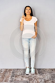 Happy curly woman having idea and showing forefinger while looking up over gray background.