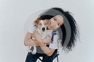 Happy curly woman tilts head, holds pedigree dog, has cheerful expression, smiles pleasantly, has curly hairstyle, wears white t