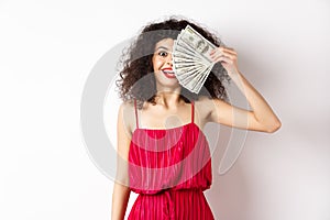 Happy curly woman winning money, holding dollar bills on half of face, smiling excited, standing in red dress on white