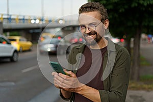 Happy customer making order for taxi cab. With smile on face, man uses phone to book ride while dusk sets in and street