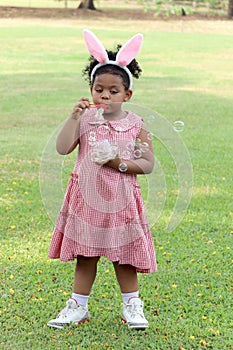 Happy cute African girl with black curly hair wearing bunny ears blowing soap bubbles in green nature garden. Kid spending time