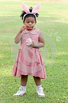 Happy cute African girl with black curly hair wearing bunny ears blowing soap bubbles in green nature garden. Kid spending time