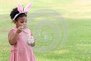 Happy cute African girl with black curly hair wearing bunny ears blowing soap bubbles in green nature garden. Kid spending time