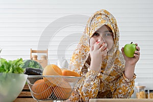 Happy cute Arab muslim kid girl wearing hijab holding and eating biting red and green apples in kitchen at home with many fruits