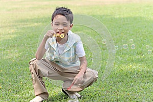 Happy cute Asian boy blowing soap bubbles while sitting on green grass in nature garden park. Kid spending time outdoors in meadow