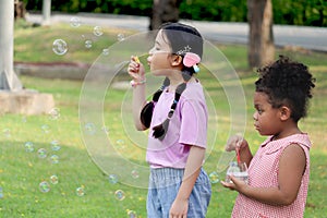 Happy cute Asian girl with curly hair African friend blowing soap bubbles in green nature garden. Kids spending time outdoor