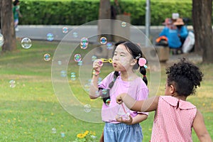 Happy cute Asian girl with curly hair African friend blowing soap bubbles in green nature garden. Kids spending time outdoor