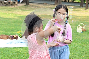 Happy cute Asian girl with curly hair African friend blowing soap bubbles in green nature garden. Kids spending time outdoor