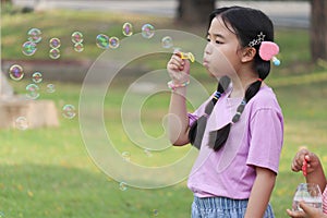 Happy cute Asian girl with pigtails blowing soap bubbles in green nature garden. Kid spending time outdoor in meadow. Cute child