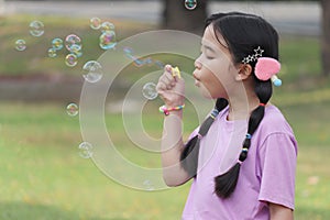 Happy cute Asian girl with pigtails blowing soap bubbles in green nature garden. Kid spending time outdoor in meadow. Cute child