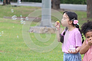 Happy cute Asian girl with pigtails blowing soap bubbles in green nature garden. Kid spending time outdoor in meadow. Cute child