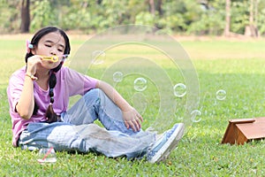Happy cute Asian girl with pigtails blowing soap bubbles while sitting on green grass in nature garden park. Kid spending time