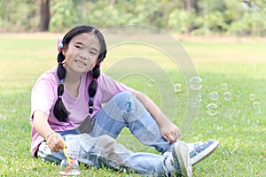 Happy cute Asian girl with pigtails blowing soap bubbles while sitting on green grass in nature garden park. Kid spending time