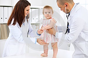 Happy cute baby at health exam at doctor`s office. Toddler girl is standing while have been keeping by two doctors
