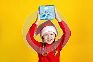 Happy cute baby holds a box over his head. Little girl in santa claus hat with christmas present on yellow background