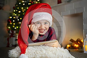 Happy cute boy in pajamas and a red hat looks at the camera while writing a letter to Santa Claus by Christmas tree in the evening