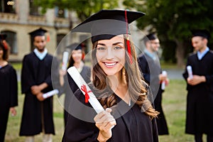 Happy cute brunette caucasian grad girl is smiling, blurred class mates are behind. She is in a black mortar board, with red tass