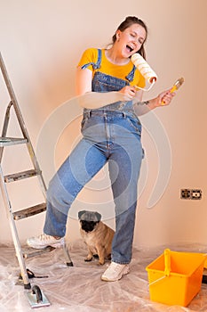 happy cute Caucasian female singing song using roller brush as a microphone. Young woman having fun during home repair
