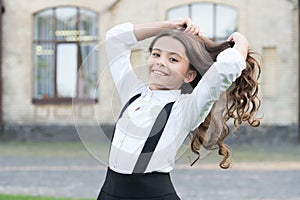 Happy cute child in school uniform fix long wavy brunette hair in schoolyard outdoors, haircare