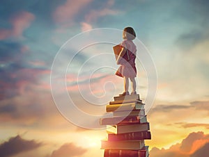 Happy cute child standing on the tower of books on background of sunset sky.