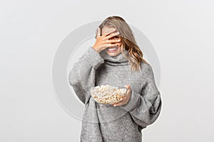 Happy cute girl with blond short hairstyle, watching movie and eating popcorn, looking through fingers at something