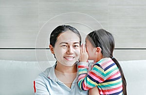 Happy cute little Asian child girl whispering a secret to her young mothers ear at home. Family and relationships concept