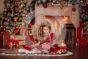 Happy cute little blonde girl with long hair in a red dress, waiting for Christmas and New year opens a gift on the