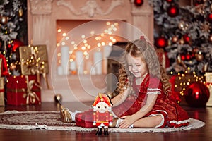 Happy cute little blonde girl with long hair in a red dress, waiting for Christmas and New year opens a gift on the