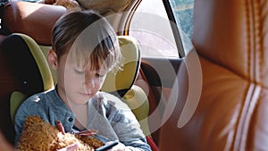 Happy cute little boy using smartphone entertainment app in car child safety seat, looking out the window on sunny day.