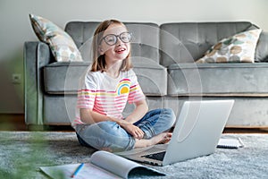 Happy cute little girl 8 years old in a striped t-shirt and jeans with glasses sits at home on a carpet in front of a laptop,