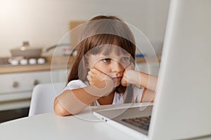 Happy cute little girl 8 years old in a striped t-shirt and jeans with glasses sits at home on a carpet in front of a laptop,