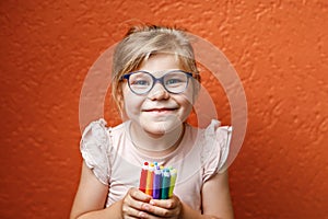 Happy cute little preschooler girl with glasses holding colorful pencils and making gesture while looking at camera