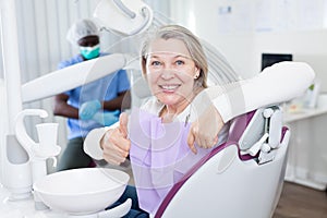 Happy cute mature woman sitting in dental chair after teeth cure giving thumb up